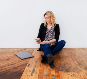 A Blonde Woman (Amy Bannon) using a cell phone with a Macbook Laptop computer beside her, sending an email about graphic design and marketing work
